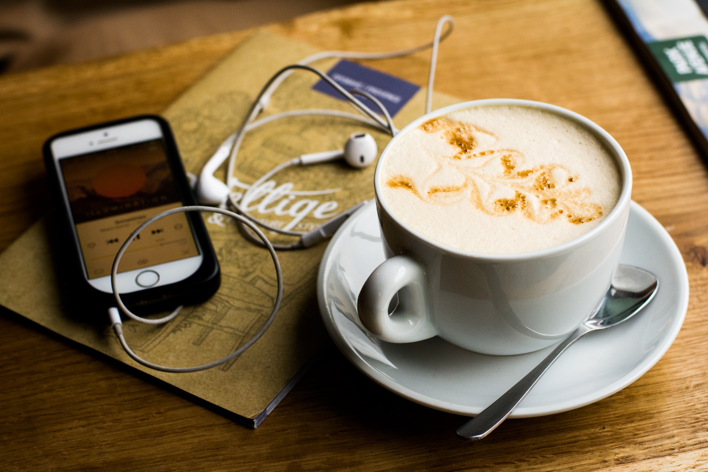 a cup of coffee on a desk alongside a mobile phone displaying a podcast on the screen with headphones plugged in on top of a brochure