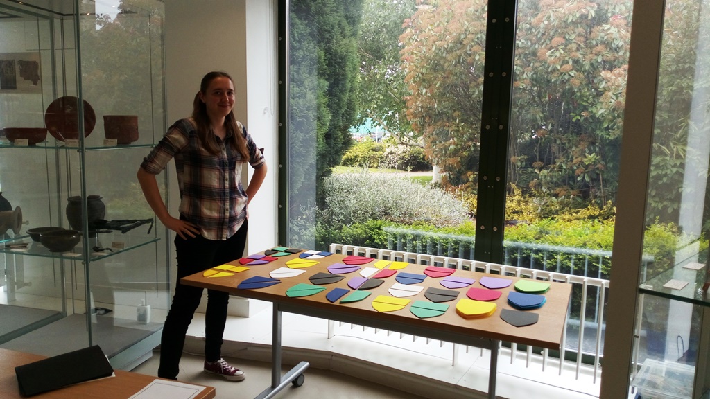 Image of a volunteer named Jasmine standing to the left of a long table. A variety of coloured shield shapes are laid out on the table top ready for use in a craft activity.